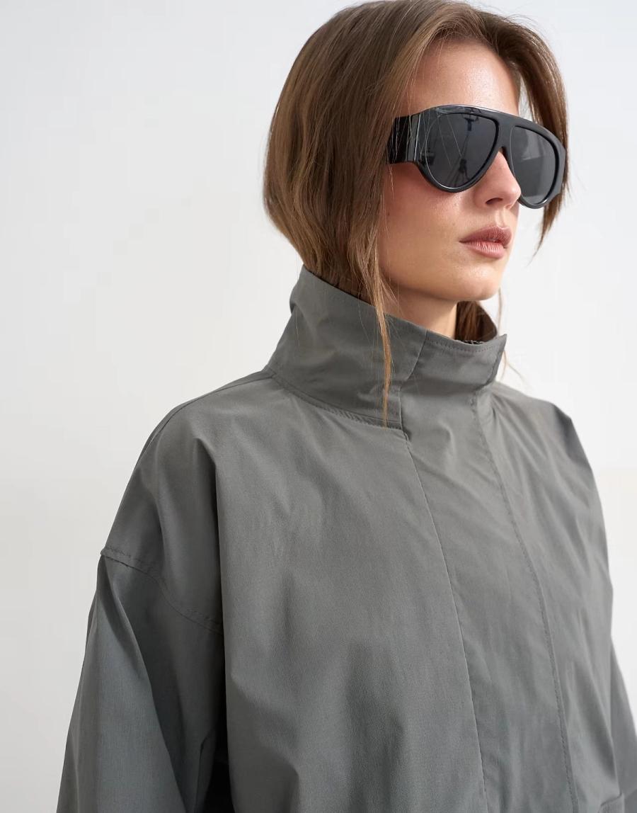 Model wearing the Technical Funnel Neck Jersey Track Top by Topshop in a stylised fashion shoot. The slate grey jacket is shown in a close-up view, highlighting the structured funnel neck, concealed front placket and relaxed shape. The model wears oversized black ski-inspired sunglasses, adding a bold fashion-led styling element to the contemporary sportswear-inspired design. The funnel neck is the main focus of the image, showing the shape and construction of the collar in detail. Iole worked as the creative pattern cutter, developing the jacket pattern and refining the proportions and construction of the funnel neck, placket and overall silhouette.