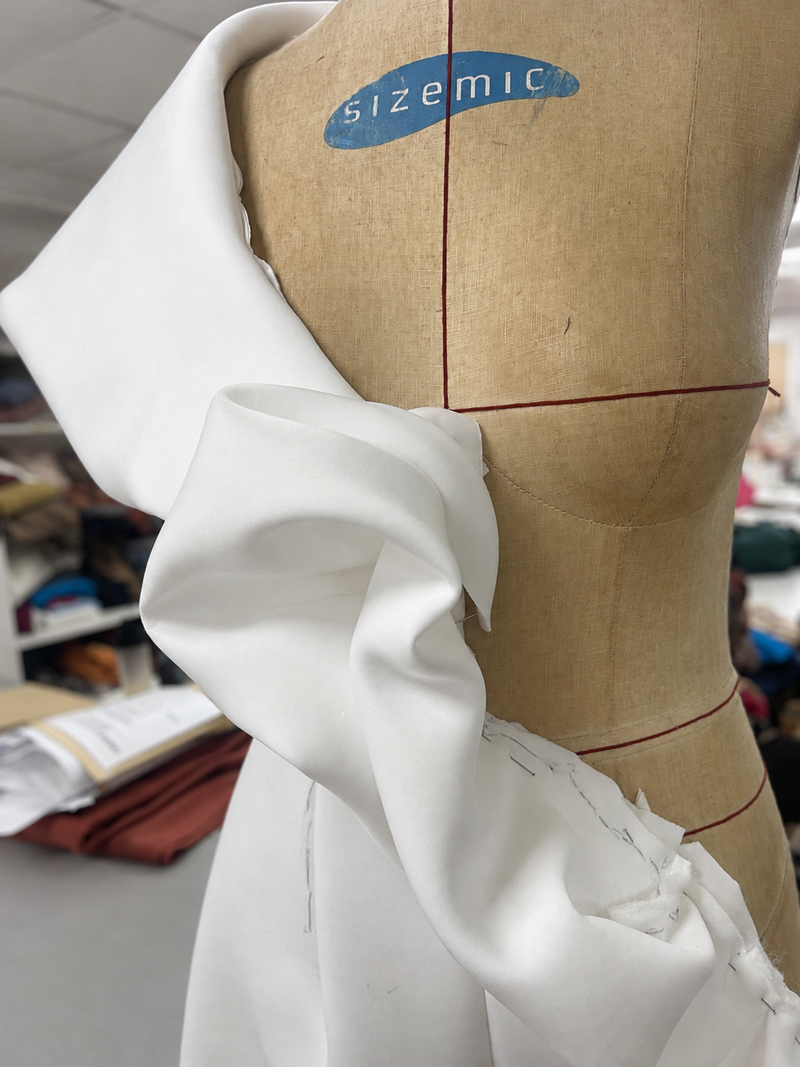 White calico toile pinned and marked with red guidelines on a dressmaker's mannequin, showing draped fabric at the shoulder.