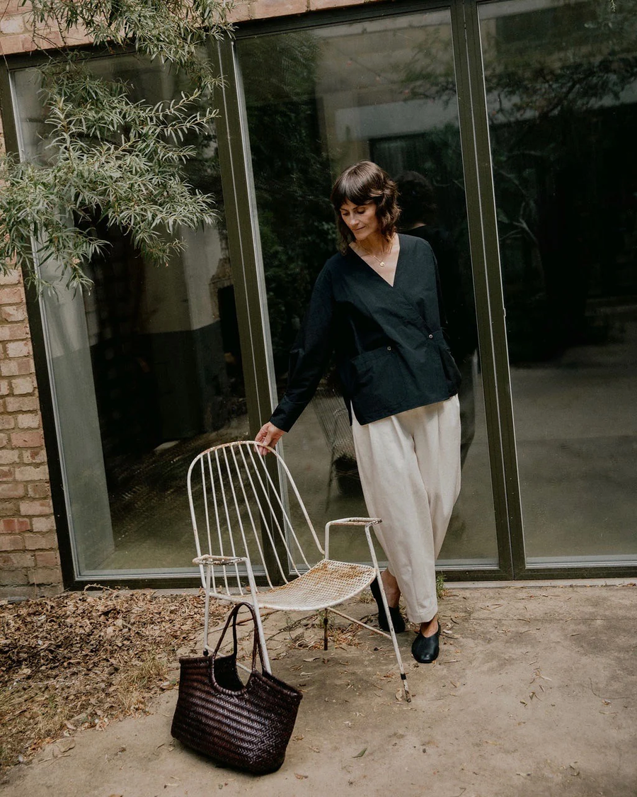 Model Niki wears the Tara Wrap Shirt in a garden setting in East London, leaning over a chair. The black organic Japanese washed cotton shirt features large patch pockets, a concealed wrap fastening that secures the pocket while also fastening the under wrap layer, and pleated cuff detailing. The relaxed silhouette combines considered construction with clever functional details. Studio Iole worked as the creative pattern cutter, developing the pattern through fittings and refinements and carefully working through the wrap front, pockets and cuffs through to the final production pattern.