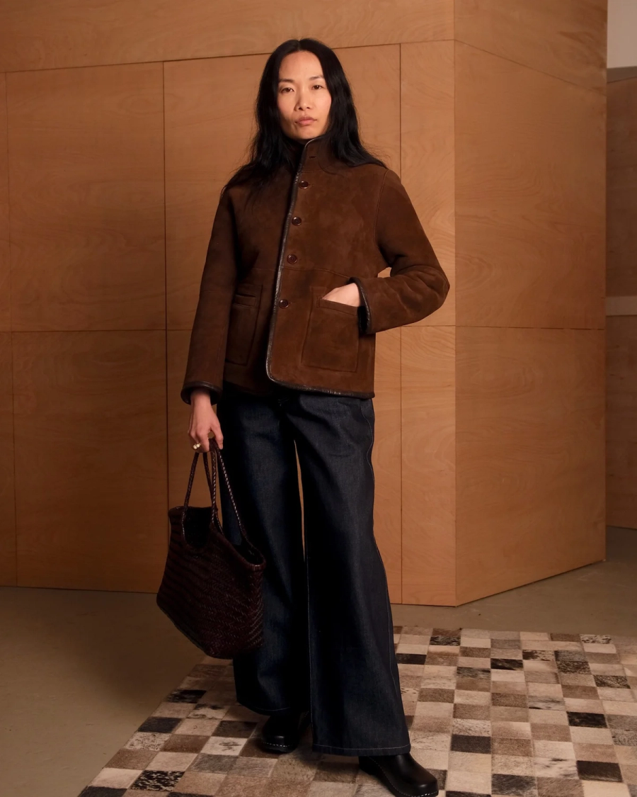Model wearing the Carolyn reversible spring weight shearling jacket by sustainable premium womenswear brand Studio Wylder. Photographed in a london studio with wood panelling as the backdrop.The jacket is crafted from lightweight shearling and features a minimal, modern silhouette designed for versatility. With 5 corzo buttons at the centre front opening and two patch pockets at the front. The hem and edging of the jacket is finished with contrast leather binding. Featuring a funnel neck collar.  Soft tailoring style.