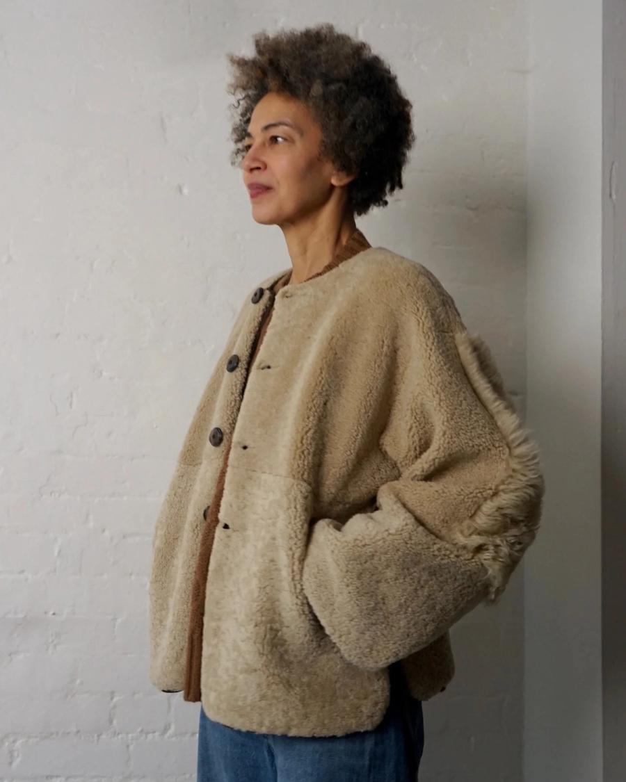 Model wearing Ara Jacket by sustainable womenswear brand Studio Wylder, handmade in London from ecru merino sheepskin. The fully reversible jacket features a round neckline, recycled horn button fastening, dropped shoulders, generous sleeves, shaped hem, fully bound edges and reversible pockets. The jacket has a relaxed, understated silhouette with a natural textured finish.