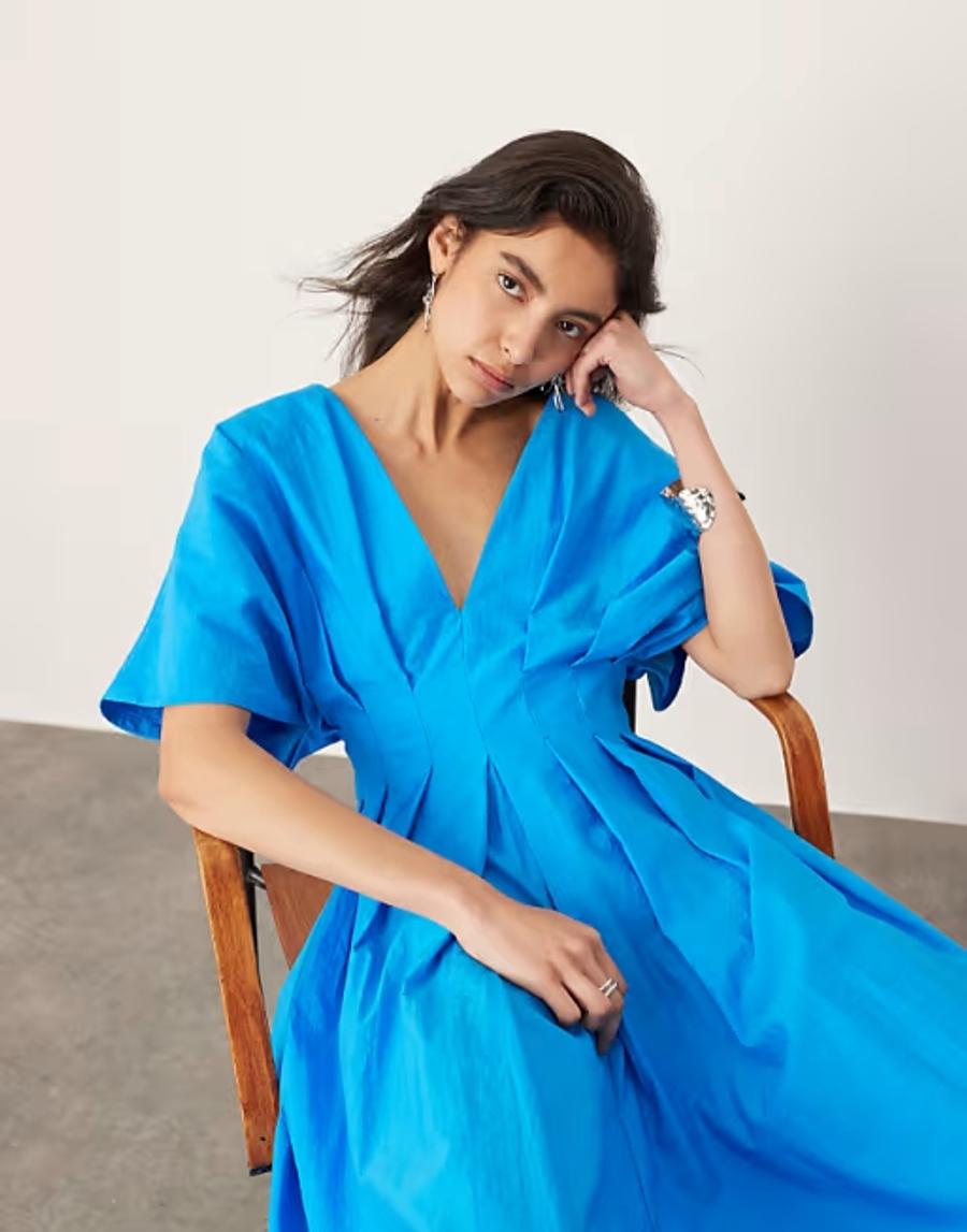 Model wearing a blue structured midi dress from ARRANGE, photographed in a professional studio setting for ASOS.com. The dress features a clean V-neckline, short sleeves, fitted bodice and defined waist, flowing into a softly structured midi-length skirt. The silhouette is fitted through the upper body before gently releasing through the skirt, highlighting the considered shaping and clean lines of the garment.