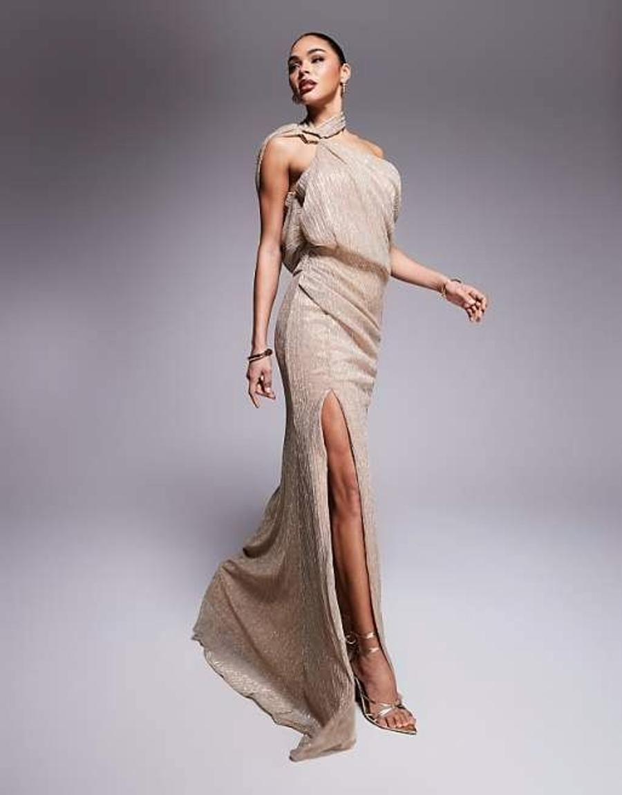 Model wearing the premium metallic one shoulder maxi dress by ASOS Design, photographed against a solid grey studio background. The gold metallic floor-length dress features an asymmetric one shoulder neckline with a gold metal trim at the right-hand side of the neck. The bodice has soft draping across the front and back, creating a wraparound cowl effect that flows around the body. The dress features a high thigh slit on the left side and a slight train that pools at the floor, creating an elongated red carpet eveningwear silhouette. The metallic fabric catches the light across the draped bodice and flowing maxi length. Iole developed the pattern as the creative pattern cutter, working through the fit, proportions and draped bodice, including developing the wraparound cowl through draping on the stand.