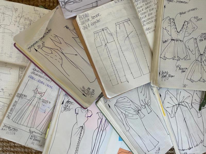 Open sketchbooks filled with fashion design sketches, technical drawings, and handwritten fabric notes.