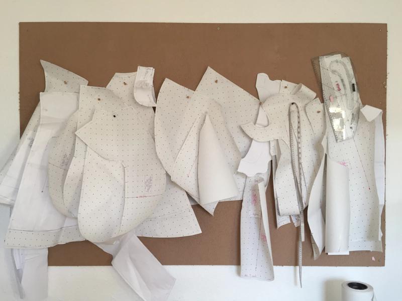 White paper pattern pieces for a garment pinned to a corkboard, showing collar, sleeve, and bodice shapes.