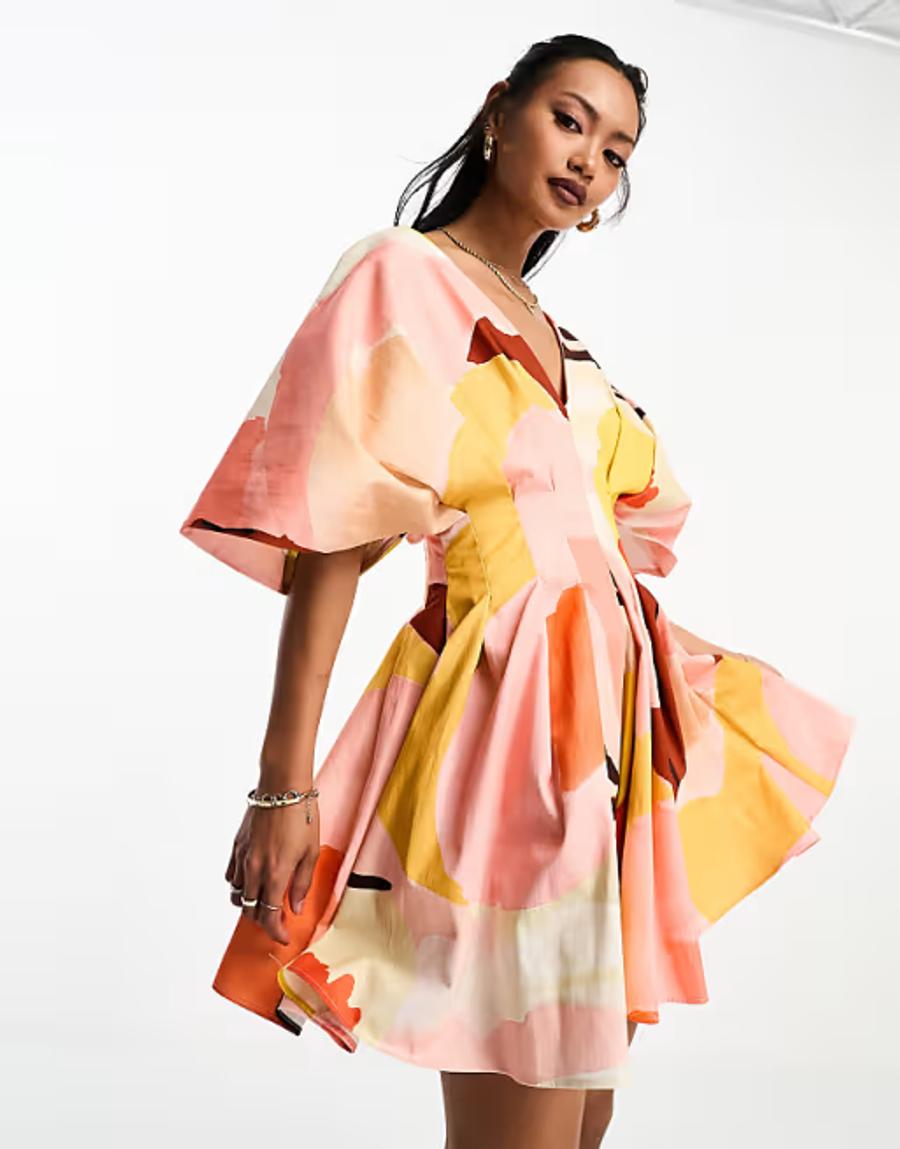 A model wears an ASOS EDITIONS structured mini dress with a bold abstract graphic print in pink, yellow, peach and orange, photographed against a plain white studio background. The dress features a V-neckline, fitted waist, voluminous short  sleeves and a structured, flared mini skirt. The pleated pattern cutting creates shape and volume through the body and skirt.