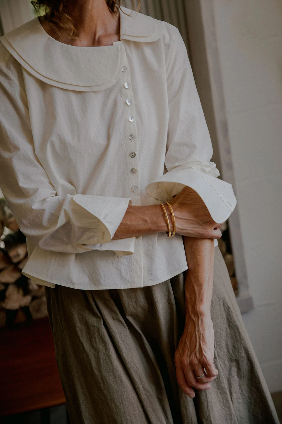 Model wearing the Nova Blouse by sustainable womenswear brand Studio Wylder, photographed in a home setting with a log pile in the background. The blouse is crafted from crisp cream Japanese 100% organic cotton and features a gentle A-line silhouette, asymmetric front button fastening, double collar and distinctive curved double cuffs, styled with the cuffs slightly scrunched up. The delicate mini mother of pearl buttons add a refined, understated luxury to the design. Studio Iole developed the creative pattern through fittings and refinements, working through to the final sealed production pattern used to produce the finished garment shown.
