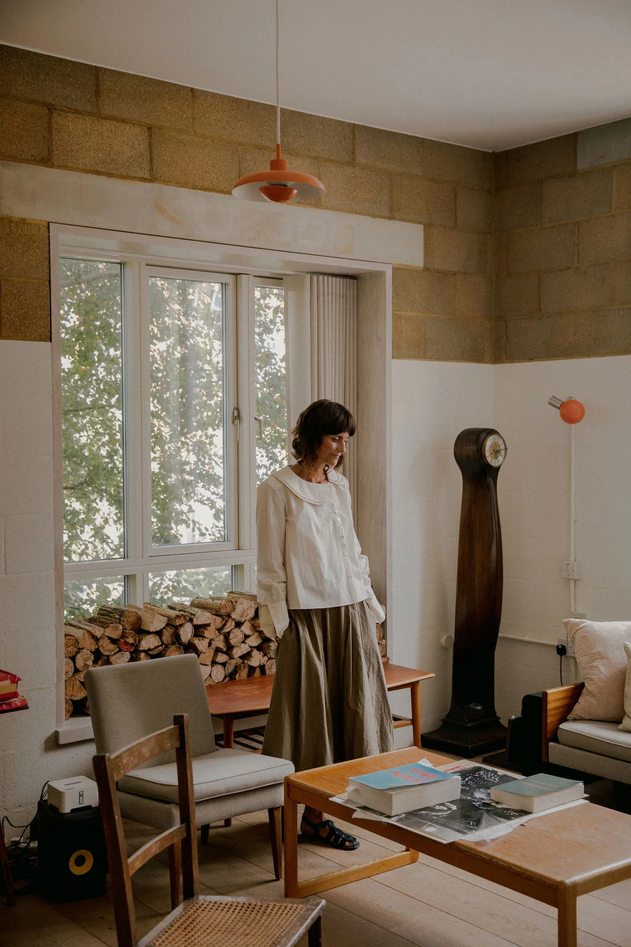 Model Nikki from Style in the Stix is wearing the Nova Blouse by sustainable womenswear brand Studio Wylder, photographed in a mid century modern home setting with a log pile in the background. The blouse is crafted from crisp cream Japanese 100% organic cotton and features a gentle A-line silhouette, asymmetric front button fastening, double collar and distinctive curved double cuffs, styled with the cuffs slightly scrunched up. The delicate mini mother of pearl buttons add a refined, understated luxury to the design. Studio Iole developed the creative pattern through fittings and refinements, working through to the final sealed production pattern used to produce the finished garment shown.