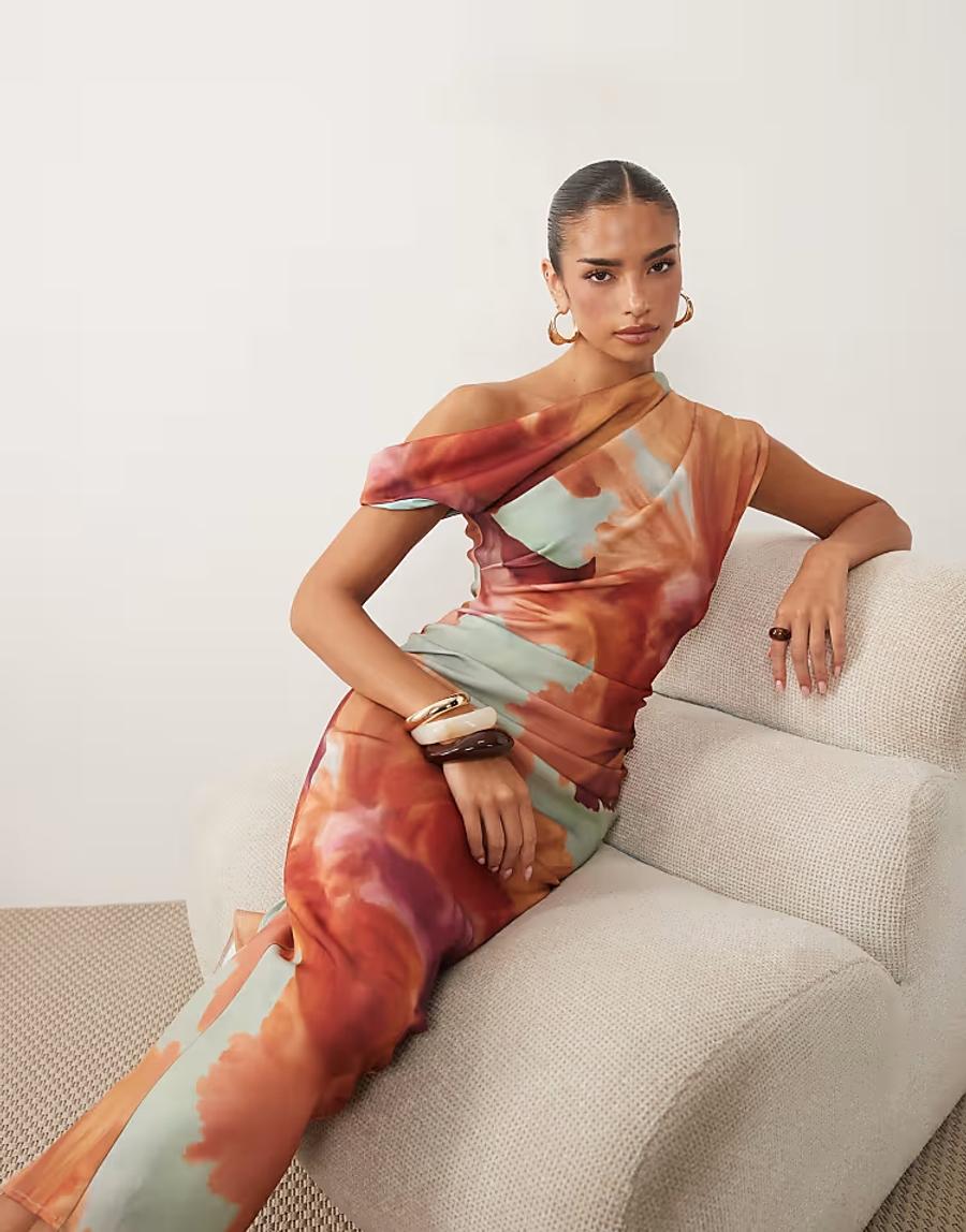 A model wears a fitted floor-length dress in an abstract watercolour print of terracotta orange, muted sage green and lavender. The dress features a dramatic asymmetric off-shoulder neckline with a draped overlay across the chest and subtle ruching through the bodice. Foam tubing is inserted into the neckline to create a raised, sculptural edge.