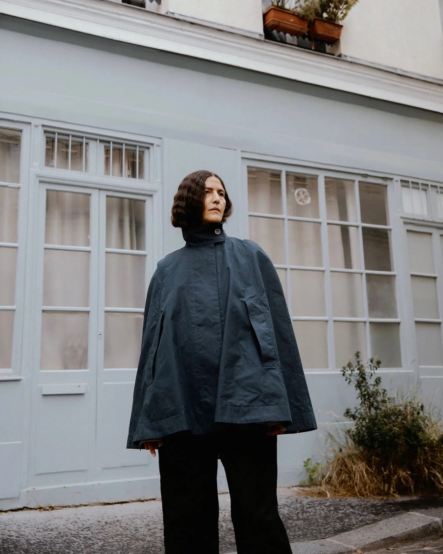 Model wearing a dark blue cape by sustainable womenswear brand Studio Wylder, photographed in the streets of Paris. The cape features a high stand collar, cape style silhouette, curved hem, structured panels and concealed pockets. Studio Iole developed the creative pattern through fittings and refinements, working through to the final sealed production pattern used to produce the finished garment shown.