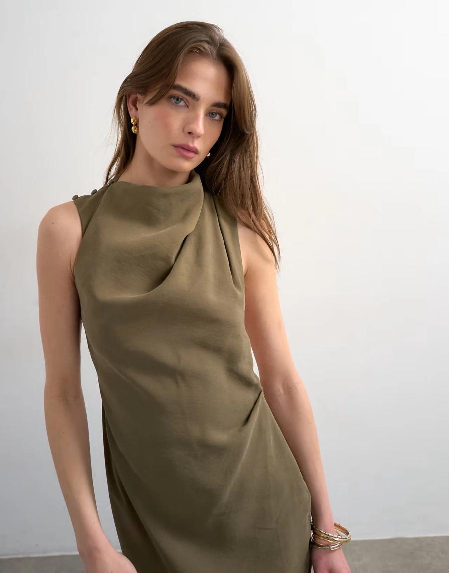 Model wearing the Khaki Modal Woven Dress in a photography studio with a concrete floor. The green woven dress has a sleeveless silhouette with a soft draped high neckline that falls around the neck and shoulder, creating gentle folds through the upper body. Pleats at the left shoulder feed into the neckline drape, while a single pleat at the left waist adds shape and a more commercial finish to the silhouette. The back features a cut-away detail that opens into the draped neckline, with a thigh-high split adding movement through the skirt. The dress is finished with three 18 ligne self-covered buttons and dye-to-match elastic loops at the right shoulder, providing an easy way into the dress while forming part of the design detail. Studio Iole developed the pattern as the creative pattern cutter, working through the drape, neckline, shoulder pleats, back cut-away and overall proportions using CLO3D mock-ups alongside the designers before moving into sampling.