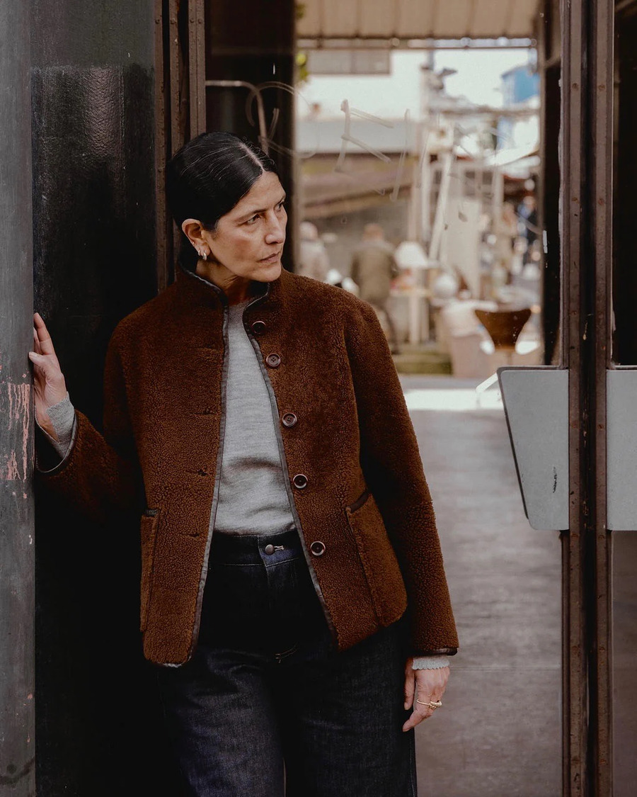 Model wearing the Carolyn reversible spring weight shearling jacket by sustainable premium womenswear brand Studio Wylder. Photographed on the streets on Paris France. The jacket is crafted from lightweight shearling and features a minimal, modern silhouette designed for versatility. with 5 corzo buttons at the centre front opening and two patch pockets at the front. The hem and edging of the jacket is finished with contrast leather binding. Featuring a funnel neck collar. 