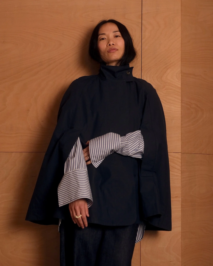 Model wearing a dark blue cape by sustainable womenswear brand Studio Wylder, photographed against a warm-toned wooden interior. The cape features a high stand collar, cape silhouette, curved hem, structured panels and concealed pockets, layered over a striped shirt. Studio Iole developed the creative pattern through fittings and refinements, working through to the final sealed production pattern used to produce the finished garment shown.