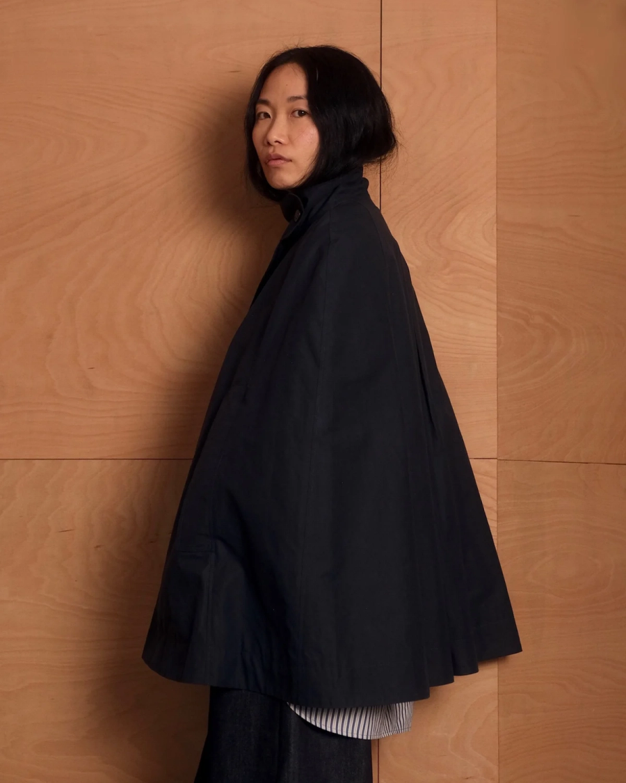 Model wearing a dark blue cape by sustainable womenswear brand Studio Wylder, photographed against a warm-toned wooden interior. The cape features a high stand collar, cape-style silhouette, curved hem, structured panels and concealed pockets, layered over a striped shirt. Studio Iole developed the creative pattern through fittings and refinements, working through to the final sealed production pattern used to produce the finished garment shown.