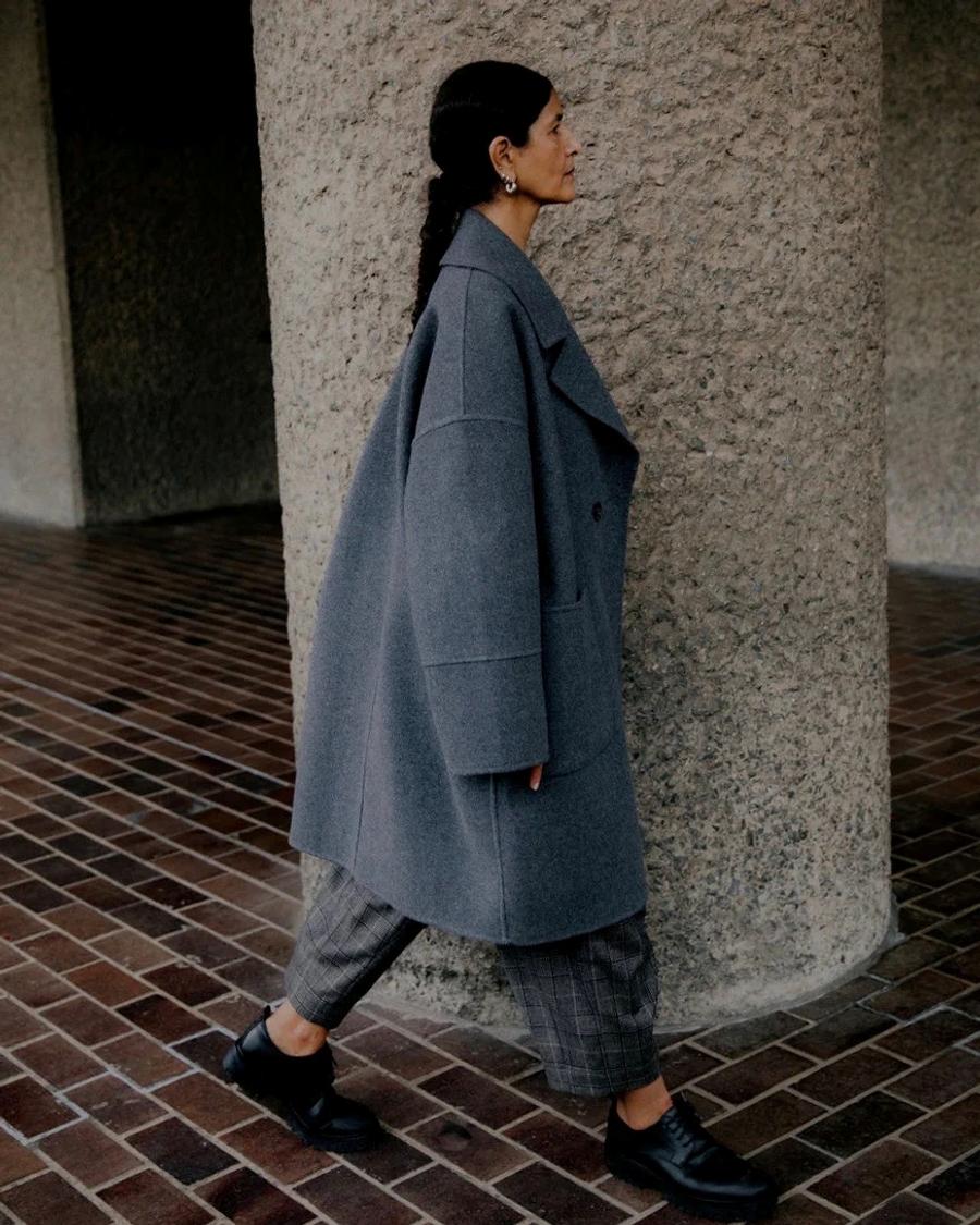 Model wearing an oversized deep grey wool coat from sustainable womenswear brand Studio Wylder’s Autumn 25 collection, photographed walking past a column at the Barbican. The coat features an oversized tailored silhouette, cocoon shaped sleeve, dropped shoulder,  structured construction and a minimalist finish. Studio Iole developed the creative pattern through fittings and refinements, working through to the final sealed production pattern used to produce the finished garment shown. Tailored Pattern Cutting.
