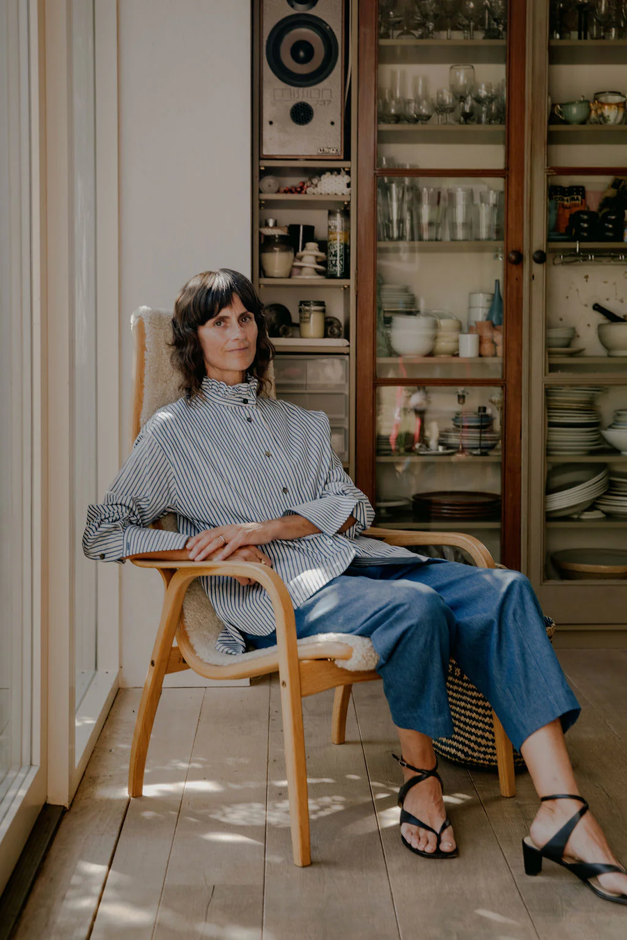 Model Nikki wearing the Victoria Ruffle Neck Blouse in a home setting in East London. The blouse features a relaxed silhouette with a soft ruffle standing at the neckline, a wide front placket with mother of pearl buttons and deep French cuffs. The organic cotton stripe fabric gives the blouse a soft, relaxed feel, while the considered cuff and placket construction add a refined finish. Studio Iole developed the pattern, refining the fit and construction through fittings through to the final production pattern.