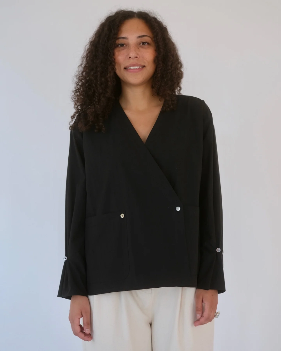 Model Niki wears the Tara Wrap Shirt in a garment studio shoot in East London. The black organic Japanese washed cotton shirt features large patch pockets, a concealed wrap fastening that secures the pocket while also fastening the under wrap layer, and pleated cuff detailing. The relaxed silhouette combines considered construction with clever functional details. Studio Iole worked as the creative pattern cutter, developing the pattern through fittings and refinements and carefully working through the wrap front, pockets and cuffs through to the final production pattern.