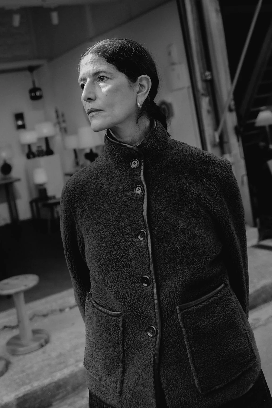 Model wearing the Carolyn reversible spring weight shearling jacket by sustainable premium womenswear brand Studio Wylder. Photographed in Black and white in front of a shop display. The jacket is crafted from lightweight shearling and features a minimal, modern silhouette designed for versatility. with 5 corzo buttons at the centre front opening and two patch pockets at the front. The hem and edging of the jacket is finished with contrast leather binding. Featuring a funnel neck collar. 