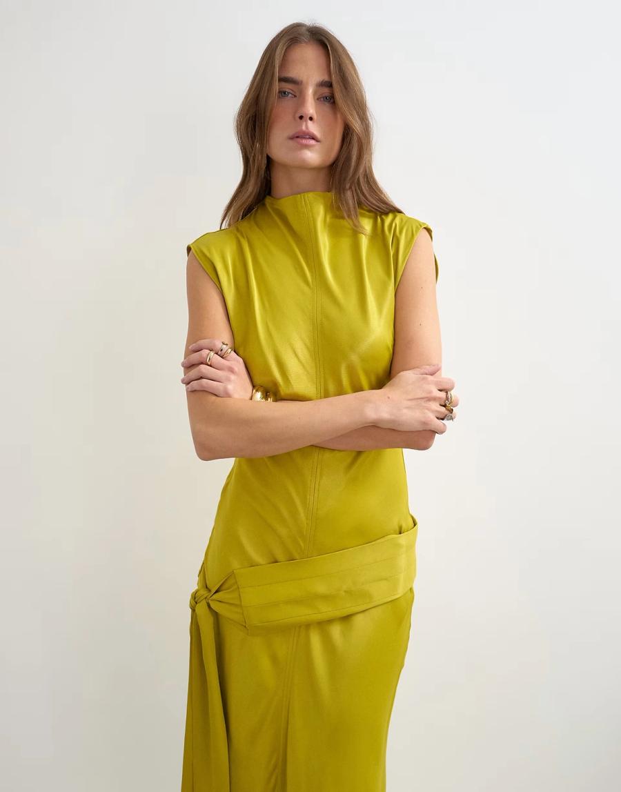 Model wearing a golden olive bias-cut satin dress in a studio setting with a concrete floor. The dress features a high neck with a slight cowl/fold at the neck with seam detailing at the centre front and centre back, which feature topstitch detailing on either side of the seam. At the dropped waist, features an Obi-style belt that goes asymmetrically across the dropped waist before tying at the wearer’s left-hand side. The back has a keyhole and button and loop closure at the neck to get into. The bias cut allows the fabric to fall naturally around the body.

Iole developed the pattern as the Creative Pattern Cutter, working through the bias-cut silhouette, fit and proportions, neckline. The fit and proportions were refined through toiling in CLO3D and draping on the stand, allowing the silhouette and construction to be checked and developed before finalising the pattern.