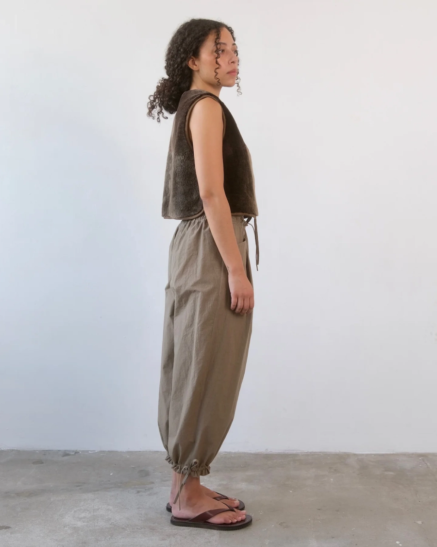 Model Tallulah wears the Tehya Drawstring Trouser in a studio in East London. Cut from crisp washed Japanese organic cotton in a rich Truffle shade, the trouser has a relaxed, gently slouching leg with a cropped hem and a shaped front created from a single pleat. The waistband is constructed with a front drawstring and softly elasticated back, allowing ease through the waist while maintaining the relaxed fit. The front pocket is engineered directly into the pleat, with the pocket opening concealed within the pleat line to keep the front clean while retaining functionality. Oversized back patch pockets are reinforced with bar tacks for stability and longevity, adding to the trouser's utilitarian feel while balancing the volume of the leg. Drawstrings at the hem allow the trouser to be worn open or gathered, changing the shape and proportion at the ankle. Studio Iole developed the pattern through fittings and refinements, working closely on the fit, pocket placement, pleat construction and adjustable details through to the final production pattern.