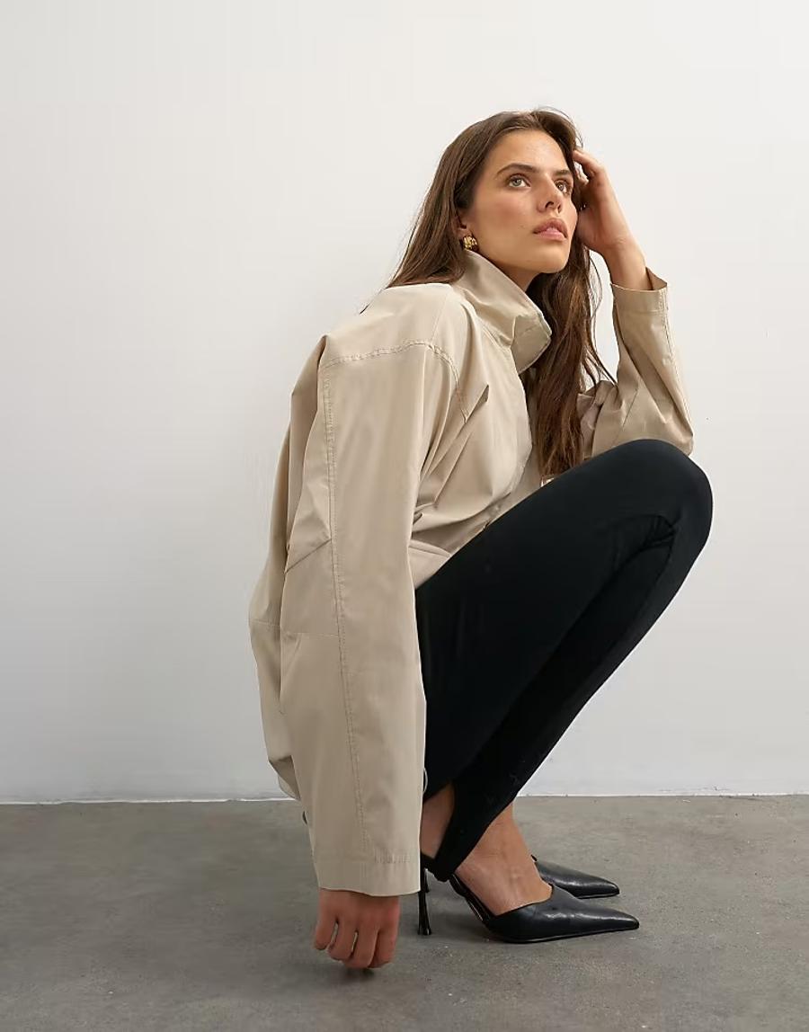 Model crouched down wearing the Technical Funnel Neck Jersey Track Top by Topshop in a stylised fashion shoot. The stone jacket is shown highlighting the structured funnel neck, concealed front placket and relaxed shape. The model wears oversized black ski-inspired sunglasses, adding a bold fashion-led styling element to the contemporary sportswear-inspired design. The funnel neck is the main focus of the image, showing the shape and construction of the collar in detail. Iole worked as the creative pattern cutter, developing the jacket pattern and refining the proportions and construction of the funnel neck, placket and overall silhouette.