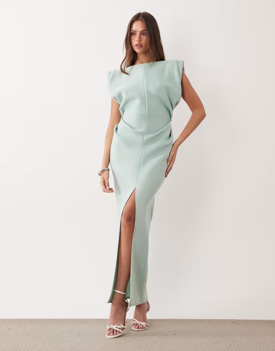 A model wears a light sage green, floor-length maxi dress against a minimalist off-white studio background. The sleeveless dress features structured pleated shoulders, a clean boat neckline and a softly draped bodice with a central seam. Sculptural pleating creates defined shaping through the bodice and shoulder, while the slim column silhouette falls to the ankles with a high front slit.