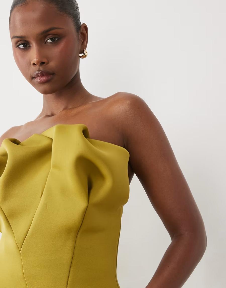 Model wearing the structured mini dress with scrunched Bardot neckline by ASOS Design, photographed against a plain off-white studio background. The avocado green scuba-style dress has a structured strapless bandeau bodice with dramatic, voluminous draping and gathered folds across the neckline created with engineered darts. The sculptural draping creates volume and texture across the upper body while the structured fabric maintains the clean shape of the mini silhouette. The model wears her dark hair pulled back into a sleek bun with small chunky gold hoop earrings. Iole developed the pattern as the creative pattern cutter, working through the fit, proportions and structured bodice, including developing the scrunched bardot neckline.
