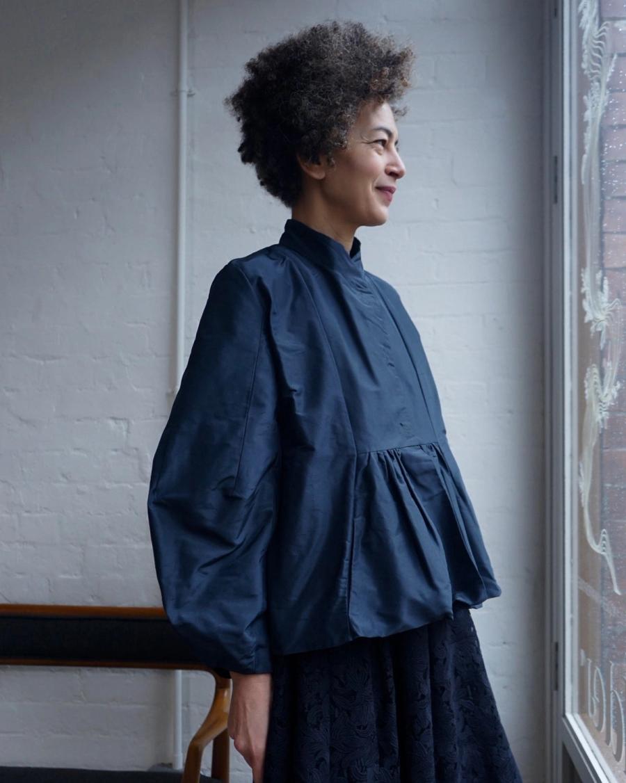 Model wearing the Alma Jacket by sustainable womenswear brand Studio Wylder. The jacket features a sculptural silhouette with a gently flared peplum, engineered sleeves, softly defined front yoke, neat mandarin collar and snap front fastening. Studio Iole developed the creative pattern through fittings and refinements, working closely on the fit, silhouette and construction through to the final sealed production pattern used to produce the finished garment shown. Made in Silk.