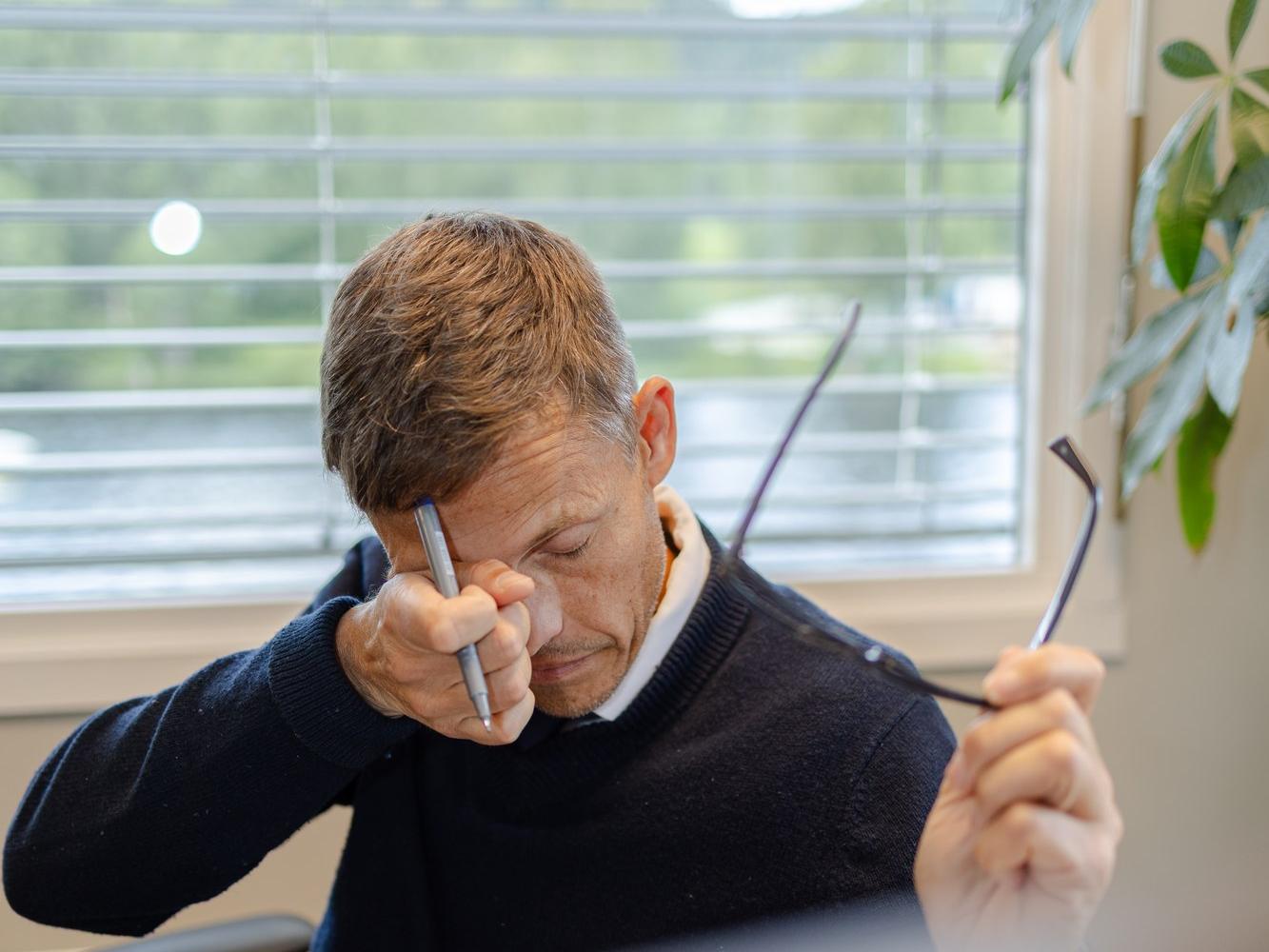 Mann med tørre og slitne øyne gnir seg i øynene ved arbeidsplassen