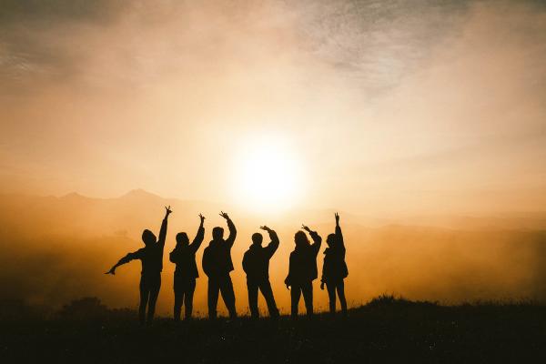 Six silhouettes of friends holding up the peace sign as the sun is rising.