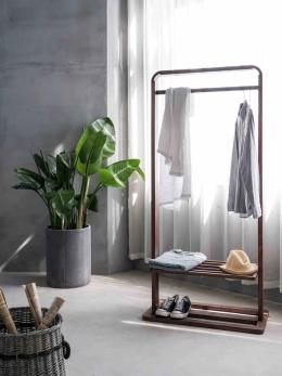 Small clothing rack with a few pieces of apparel in a large grey room with a plant next to it.