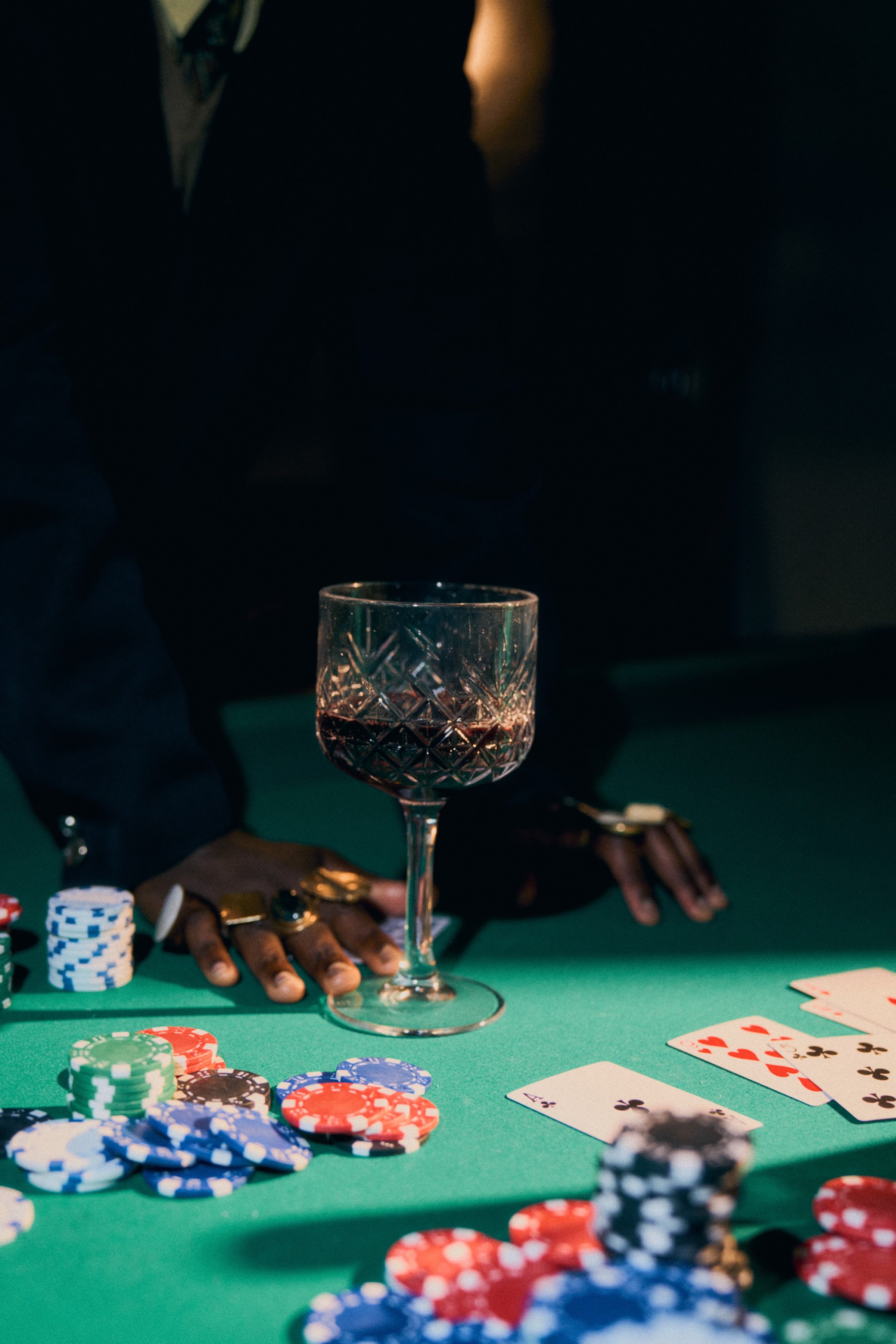 A crystal glass of red wine on a poker table, hands resting beside stacks of chips.
