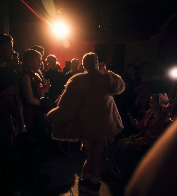 A guest in a pale fur coat walks through a party in a dark room.