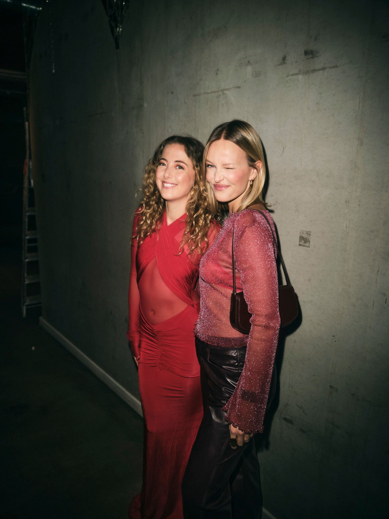 Two women in red at a party, standing against a concrete wall.