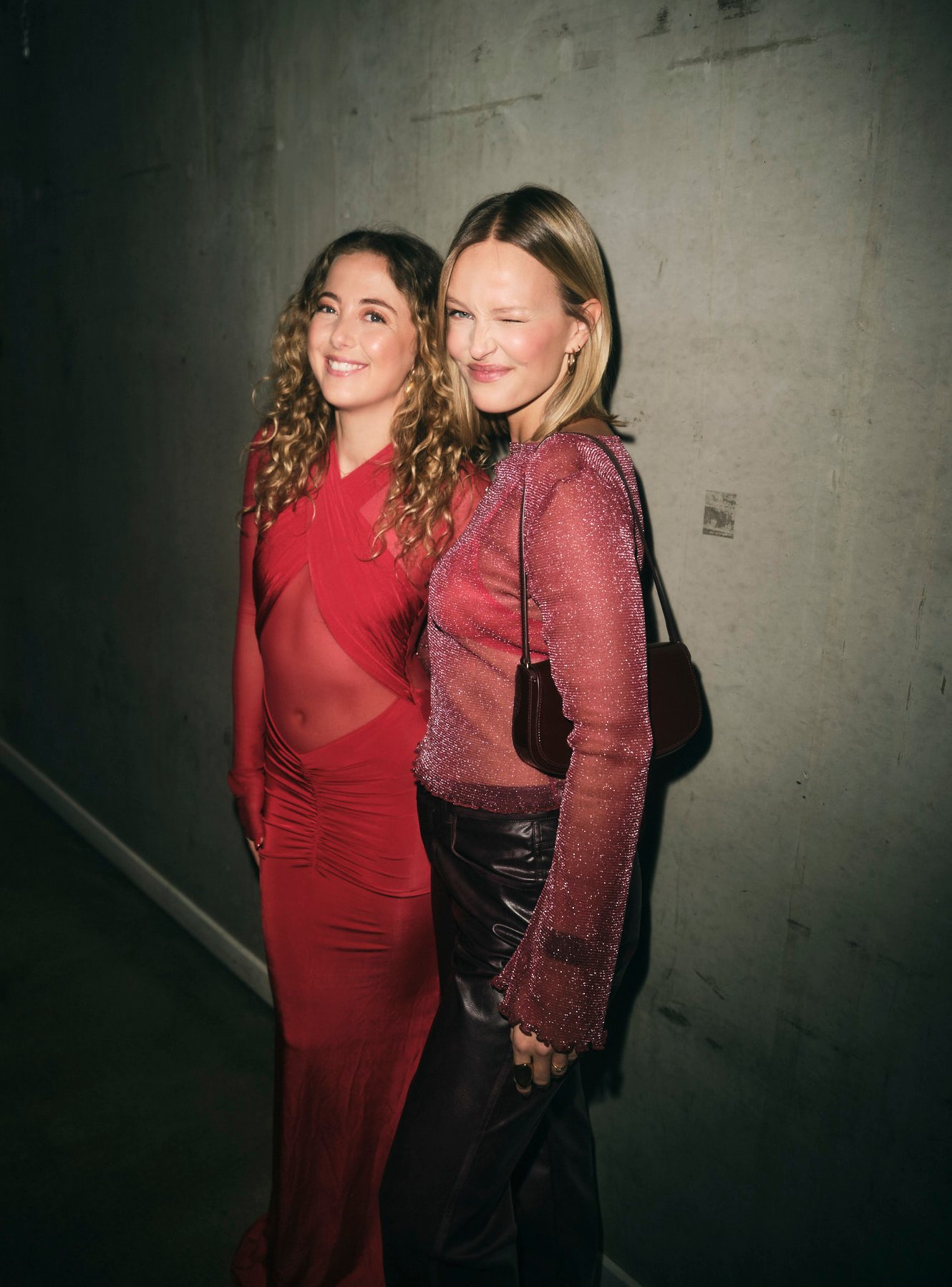 Two women in red at a party, standing against a concrete wall.