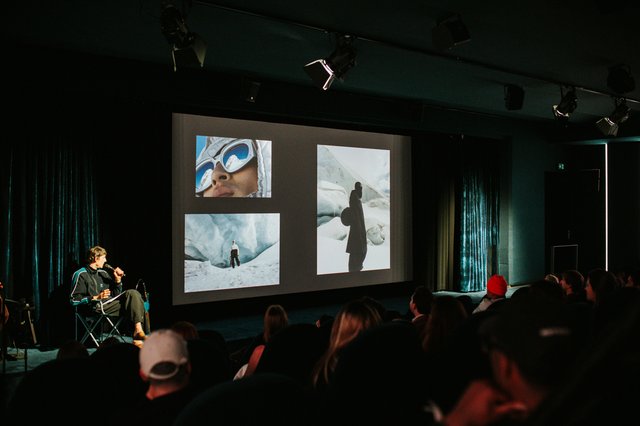 A speaker on stage in front of a screen of climbing photographs, watched by a full room.