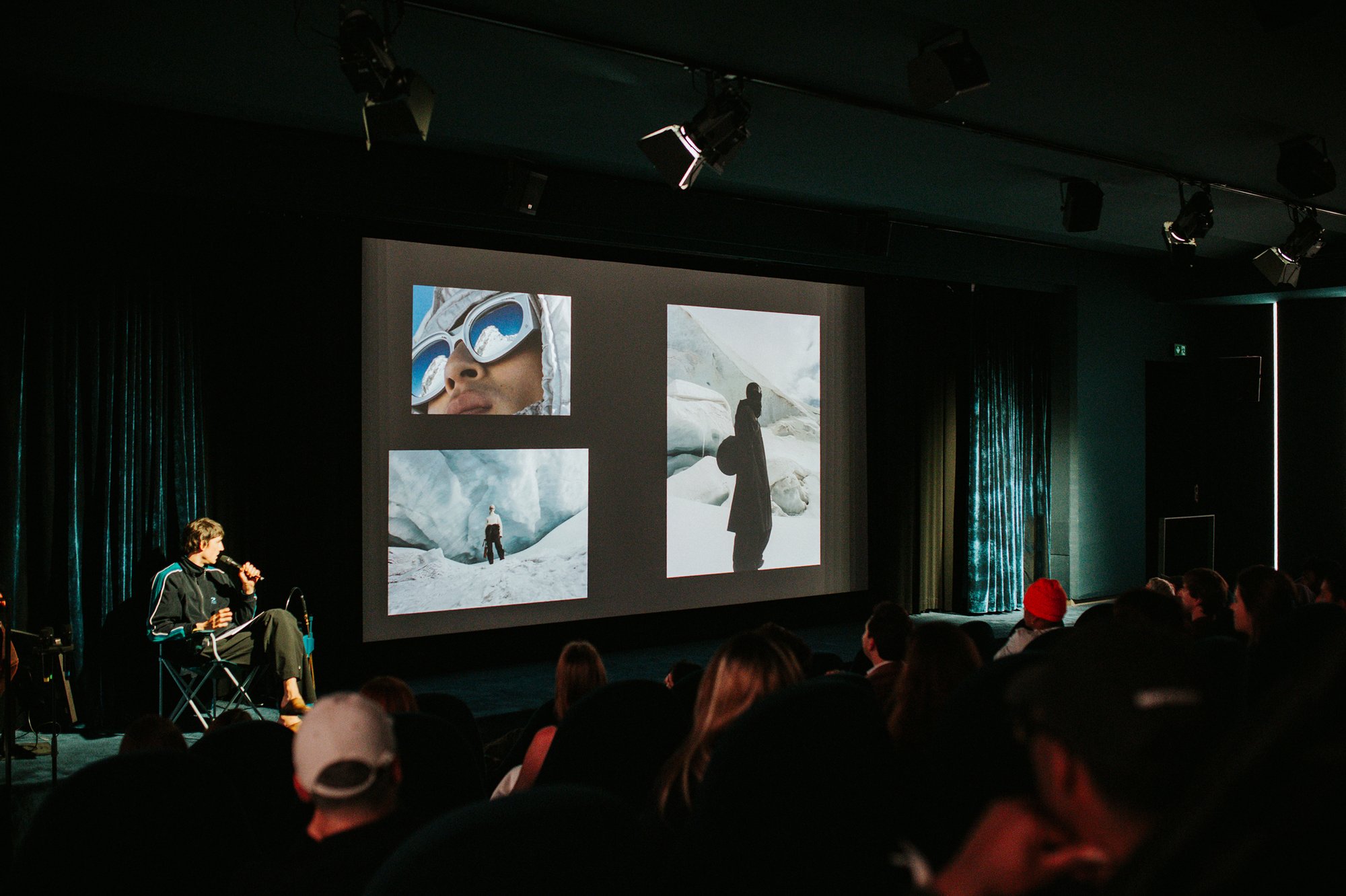 A speaker on stage in front of a screen of climbing photographs, watched by a full room.