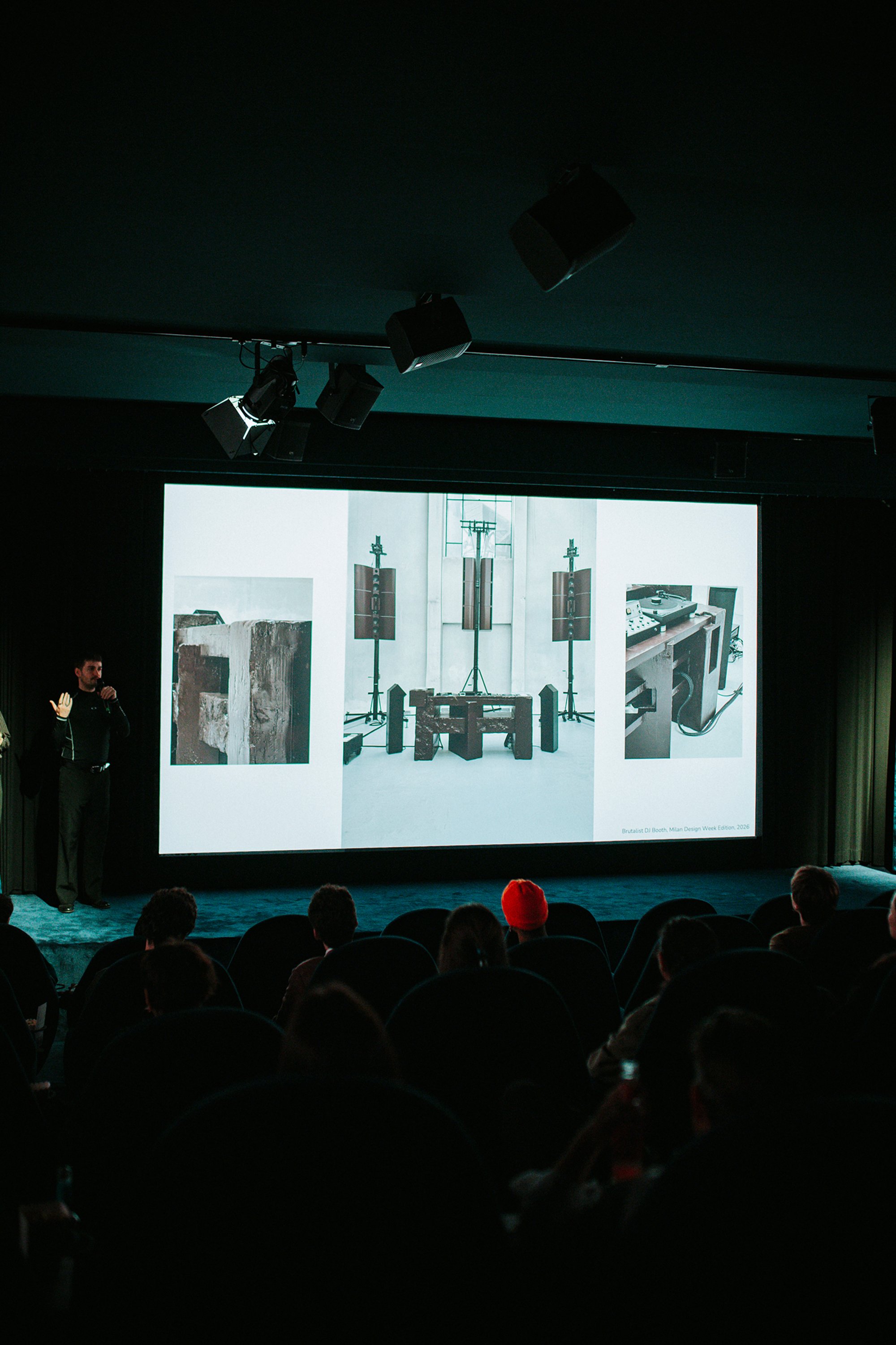 A speaker stands beside a screen showing photographs of an installation.