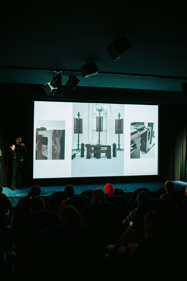 A speaker stands beside a screen showing photographs of an installation.