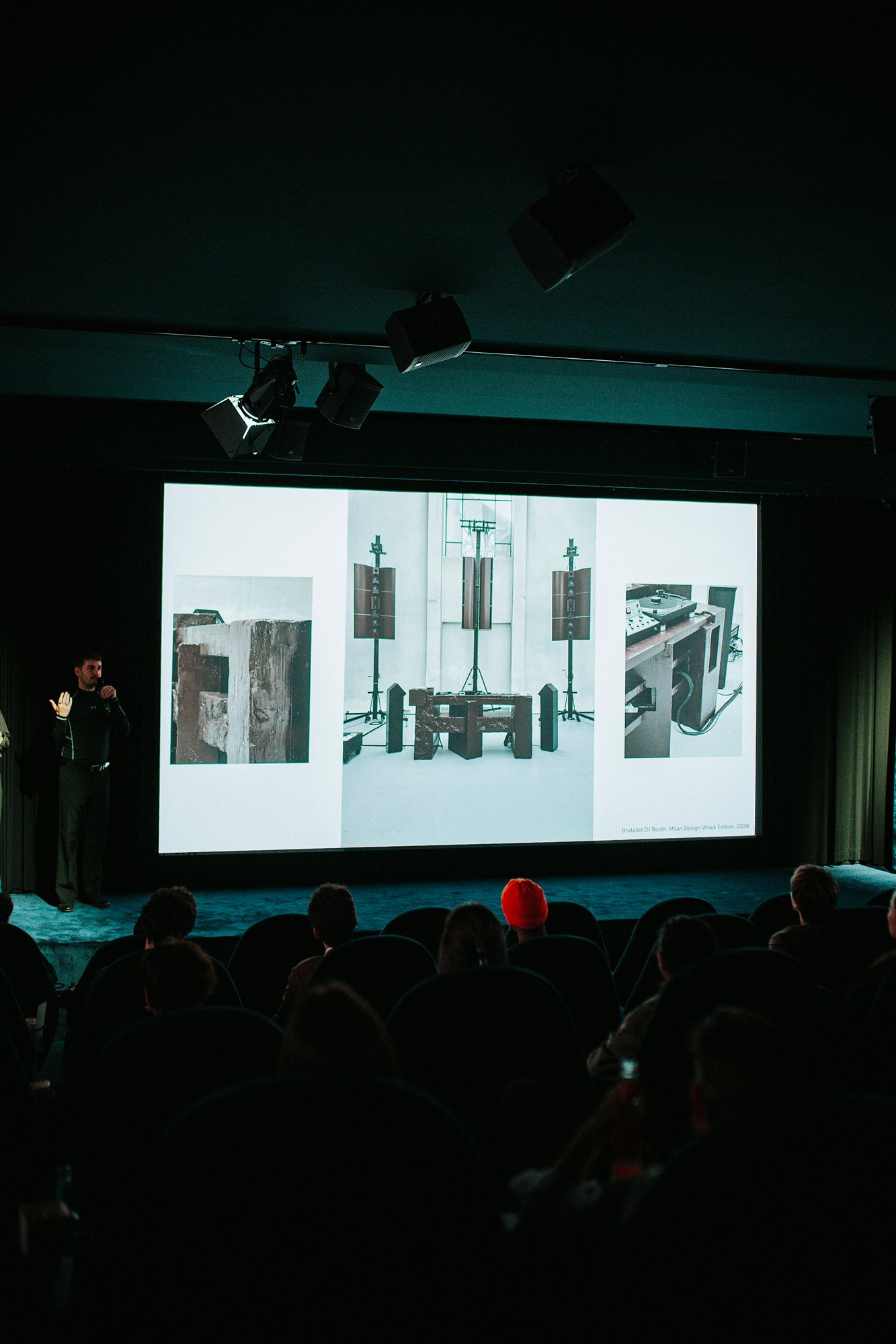 A speaker stands beside a screen showing photographs of an installation.