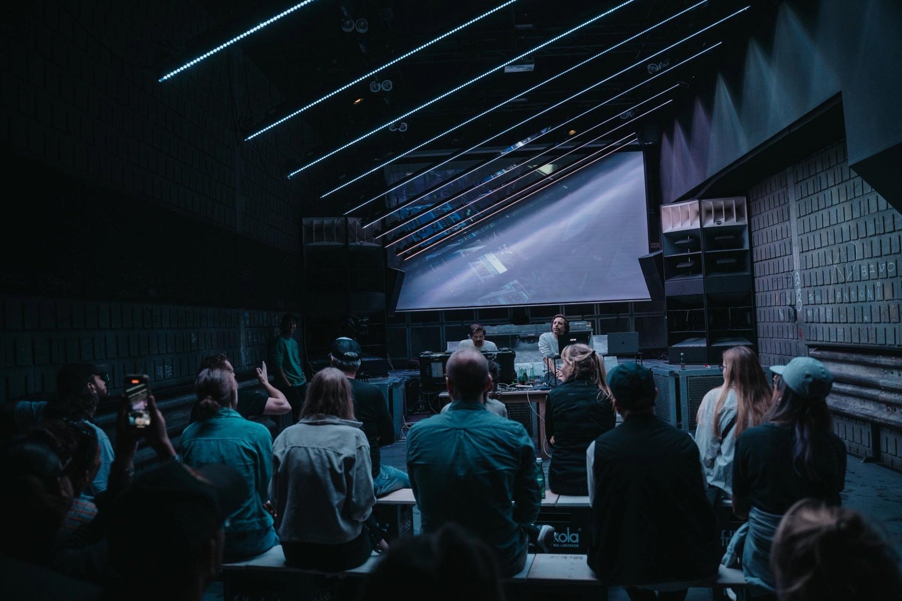 A crowd stands in a dark hall in front of a wide projection.