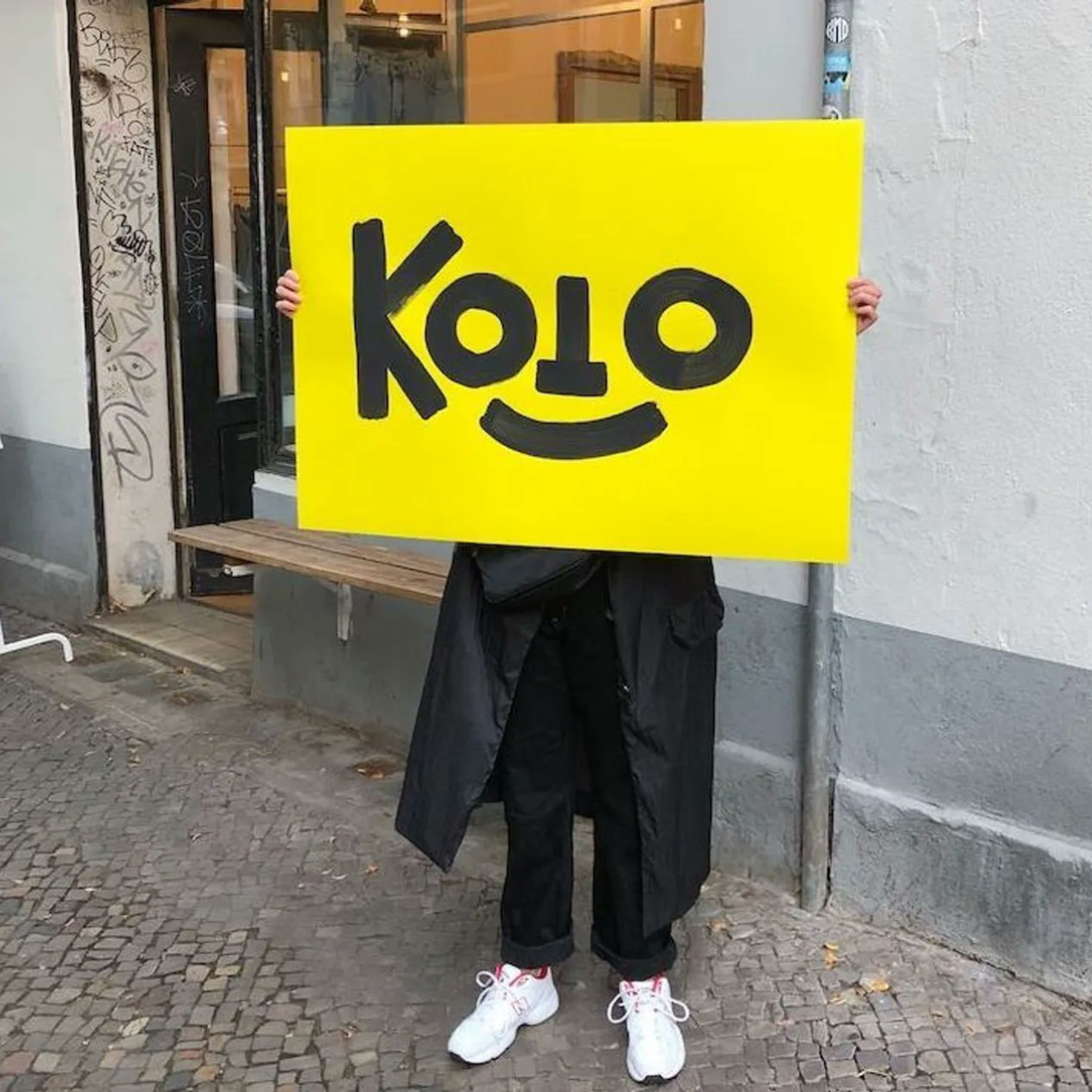 A person on the street holds a yellow Koto sign in front of their face.