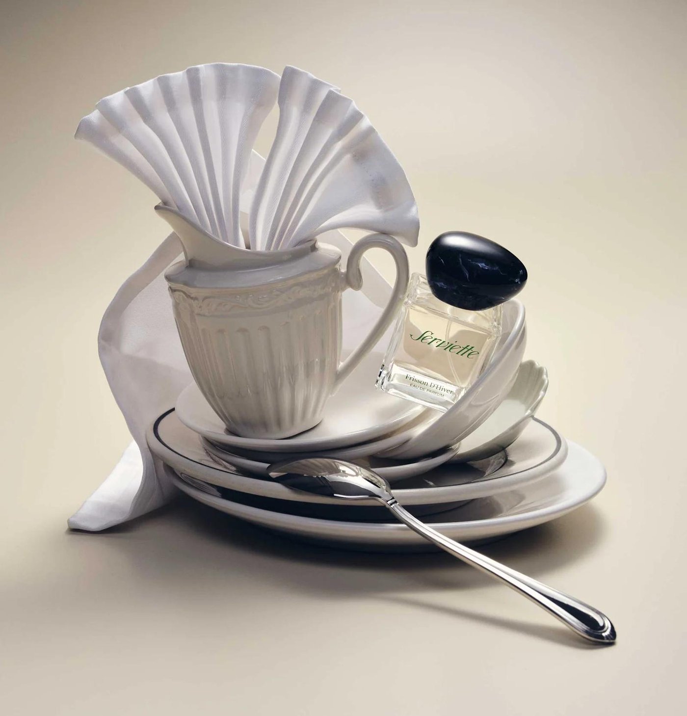 A white porcelain jug and a folded napkin stacked on plates, with a perfume bottle beside them.