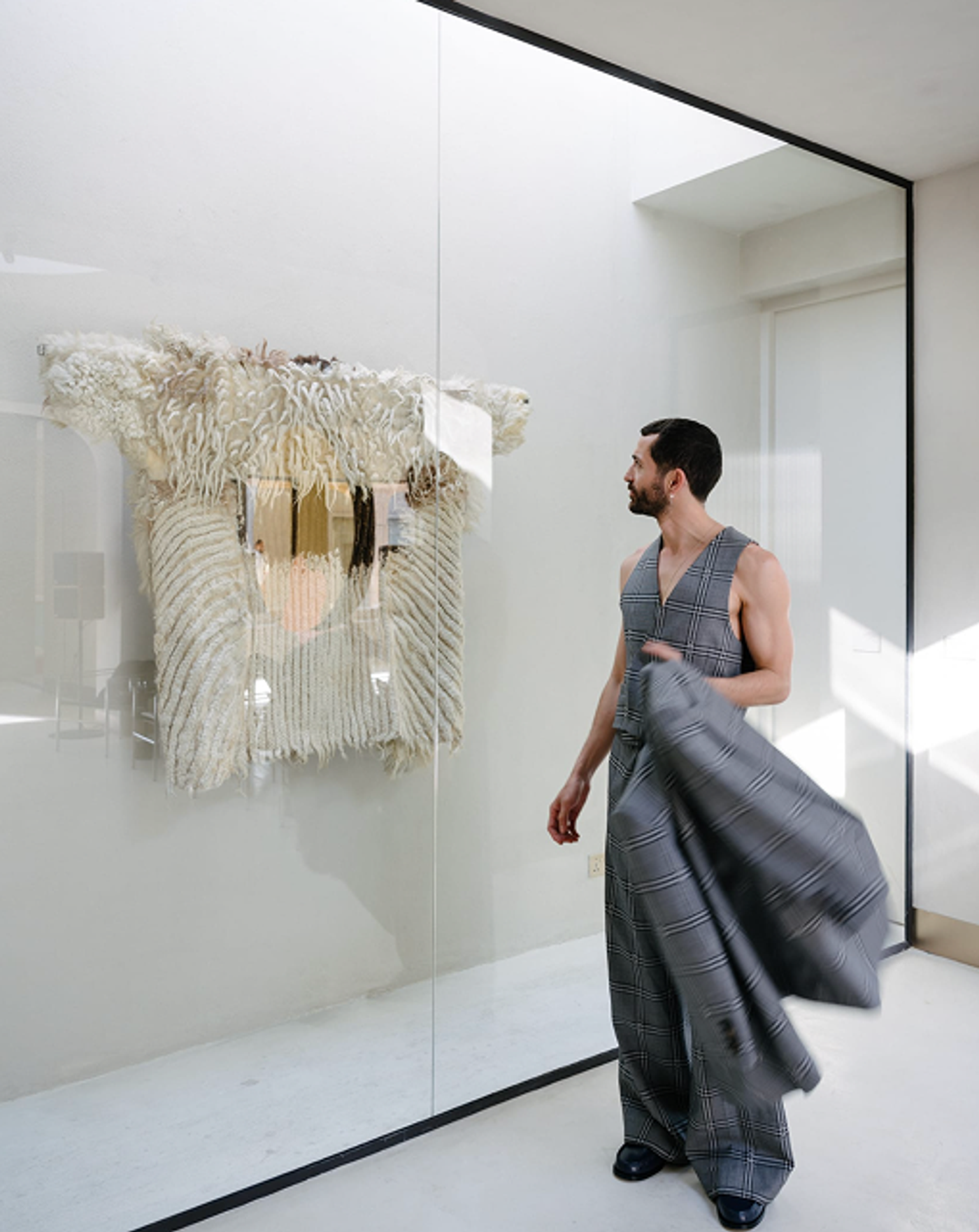 A man carries a checked coat past a woven wall hanging in a white room.