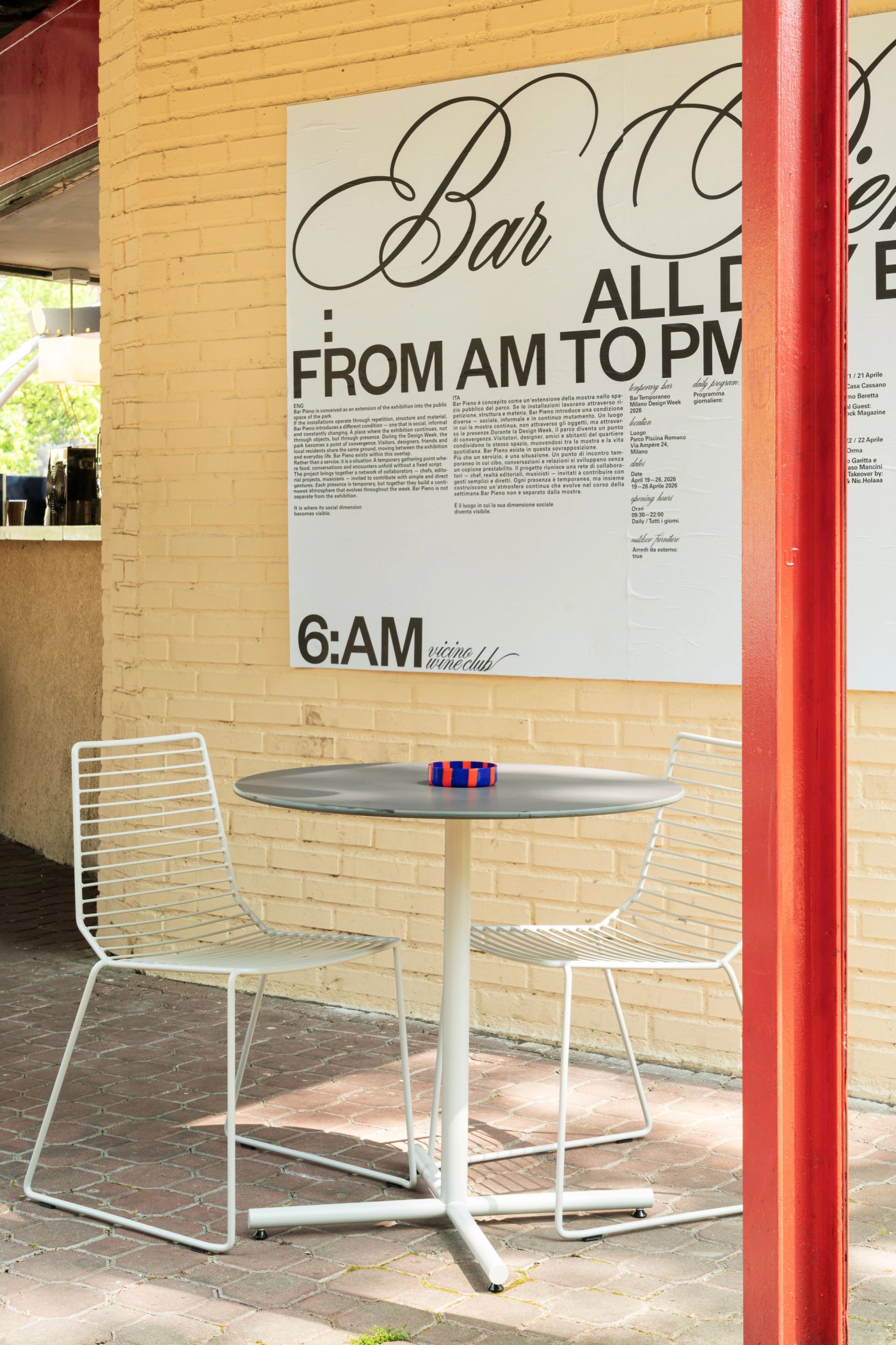 A poster for the Bar Basso collaboration on a yellow brick wall, with two wire chairs and a table under it.