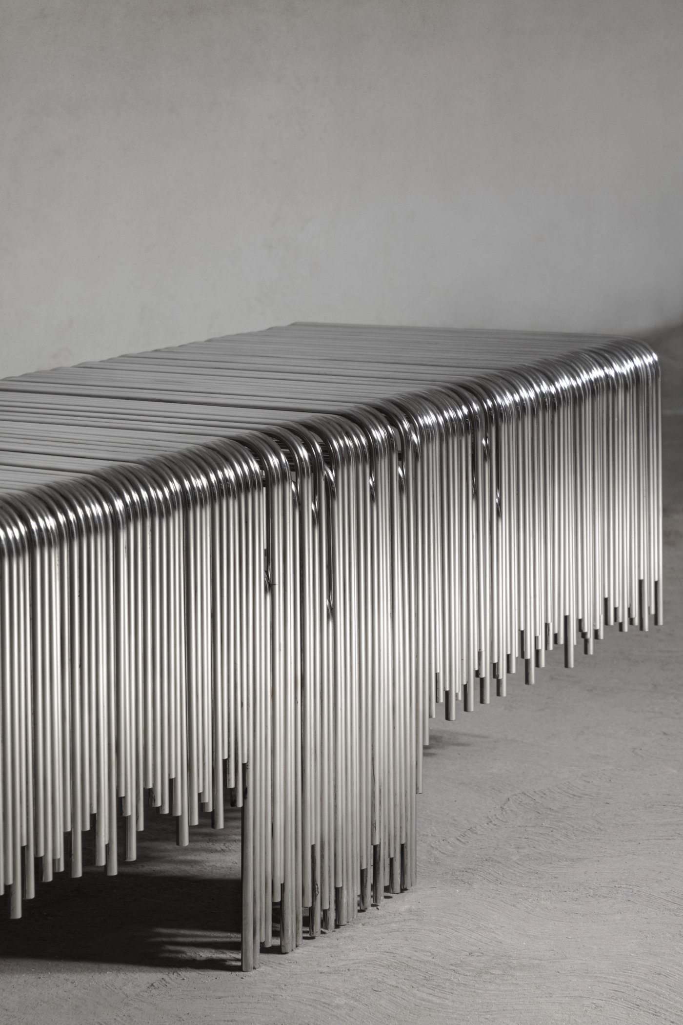 A bench made of many steel rods, standing against a plain wall.