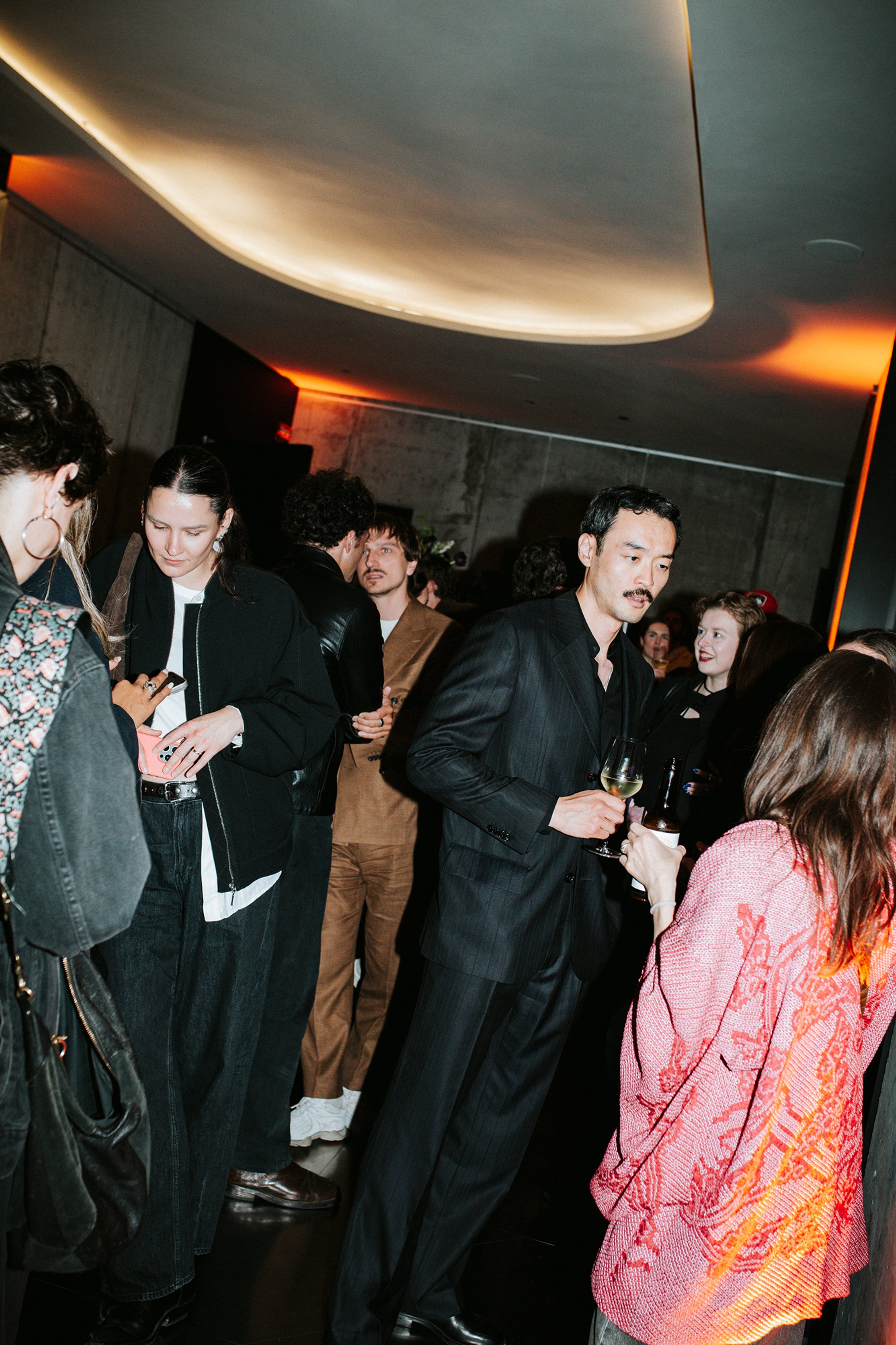 Guests with drinks at a party, standing under a lit ceiling.