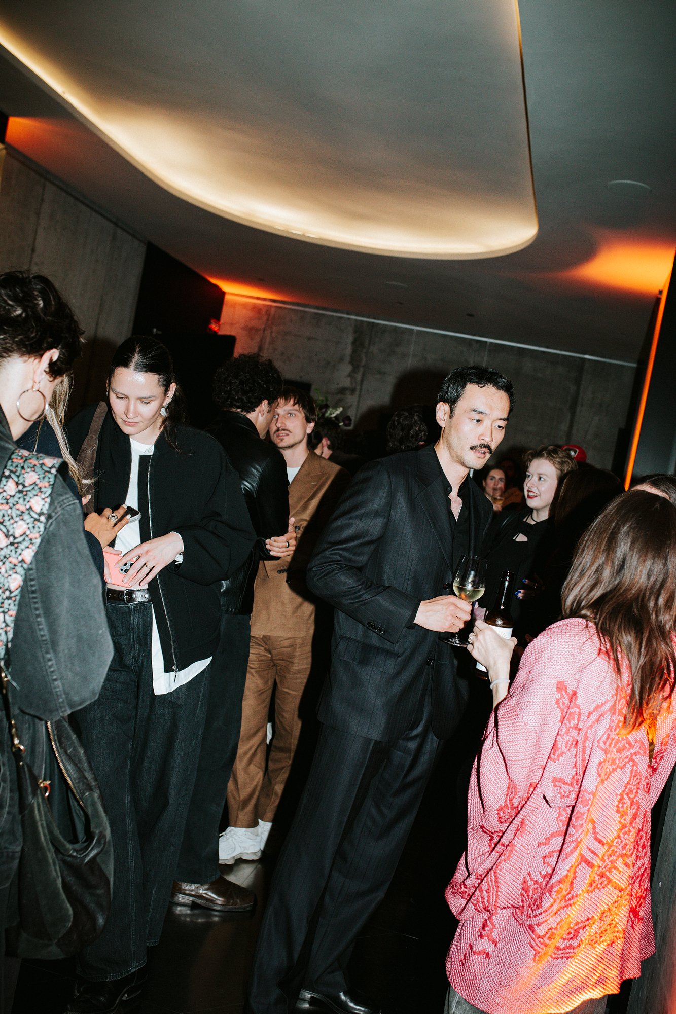 Guests with drinks at a party, standing under a lit ceiling.