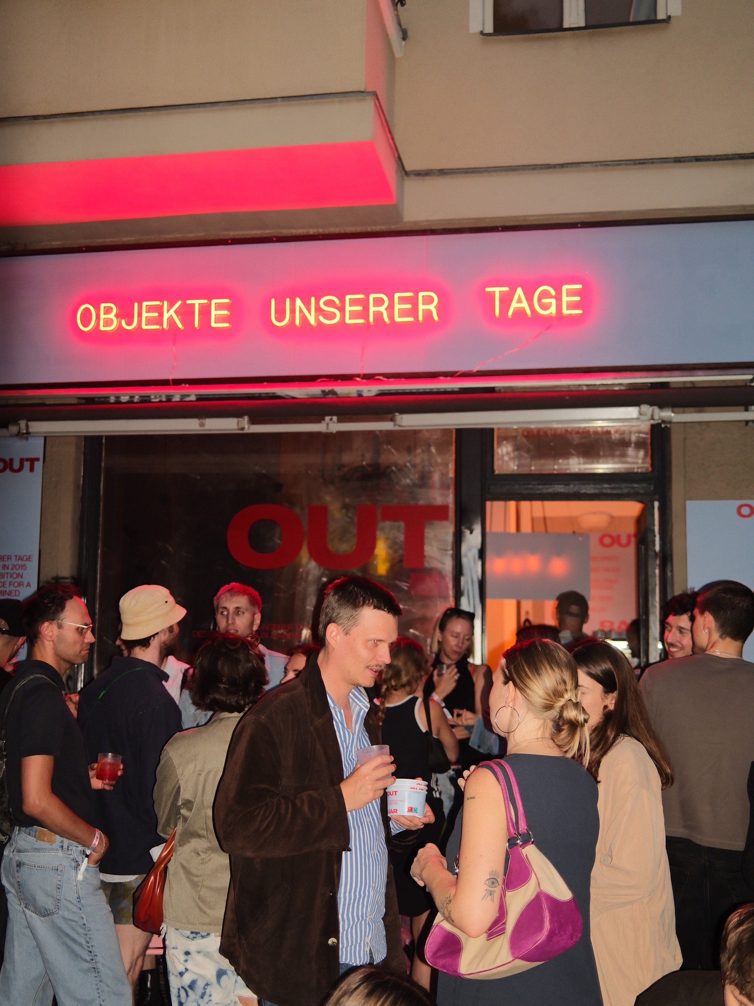 A crowd gathers outside a shopfront lit by a red neon sign reading Objekte unserer Tage.