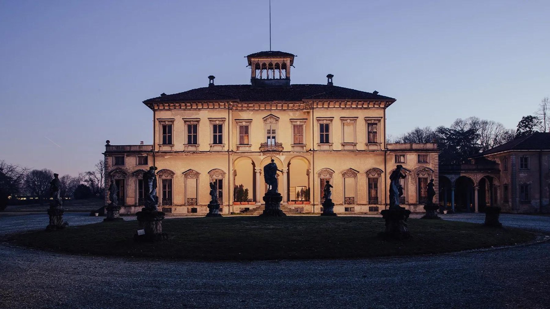 A villa lit at dusk, with statues standing along the garden in front of it.